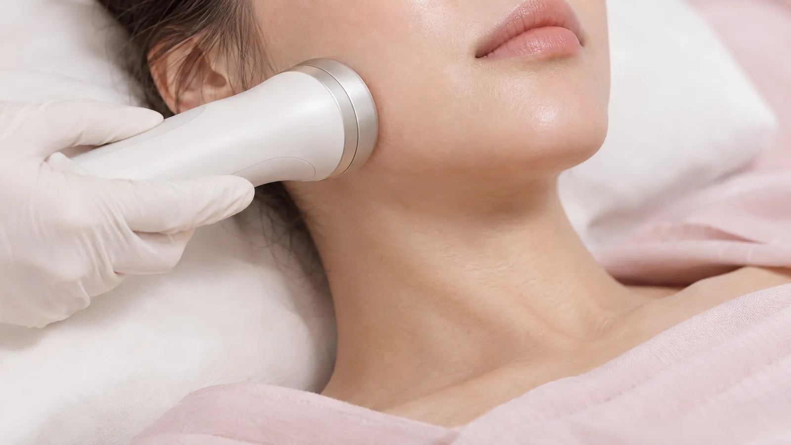 A gloved hand holds a thermal facial handpiece against an anonymous client's lower cheek beside the jawline.