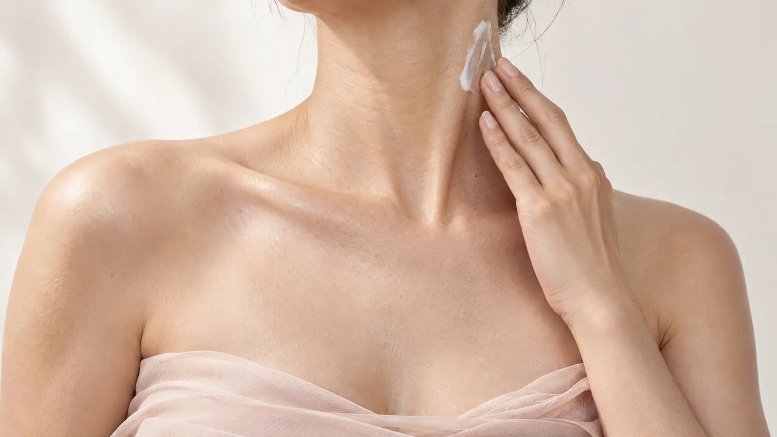 A cropped close-up shows a woman gently applying moisturizer to her neck above her collarbones.