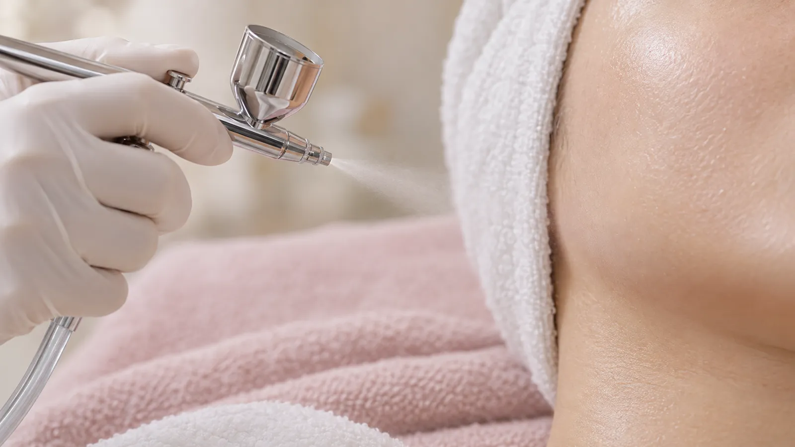 A gloved hand holds an airbrush-style facial applicator near a cropped cheek in a softly lit skincare setting.